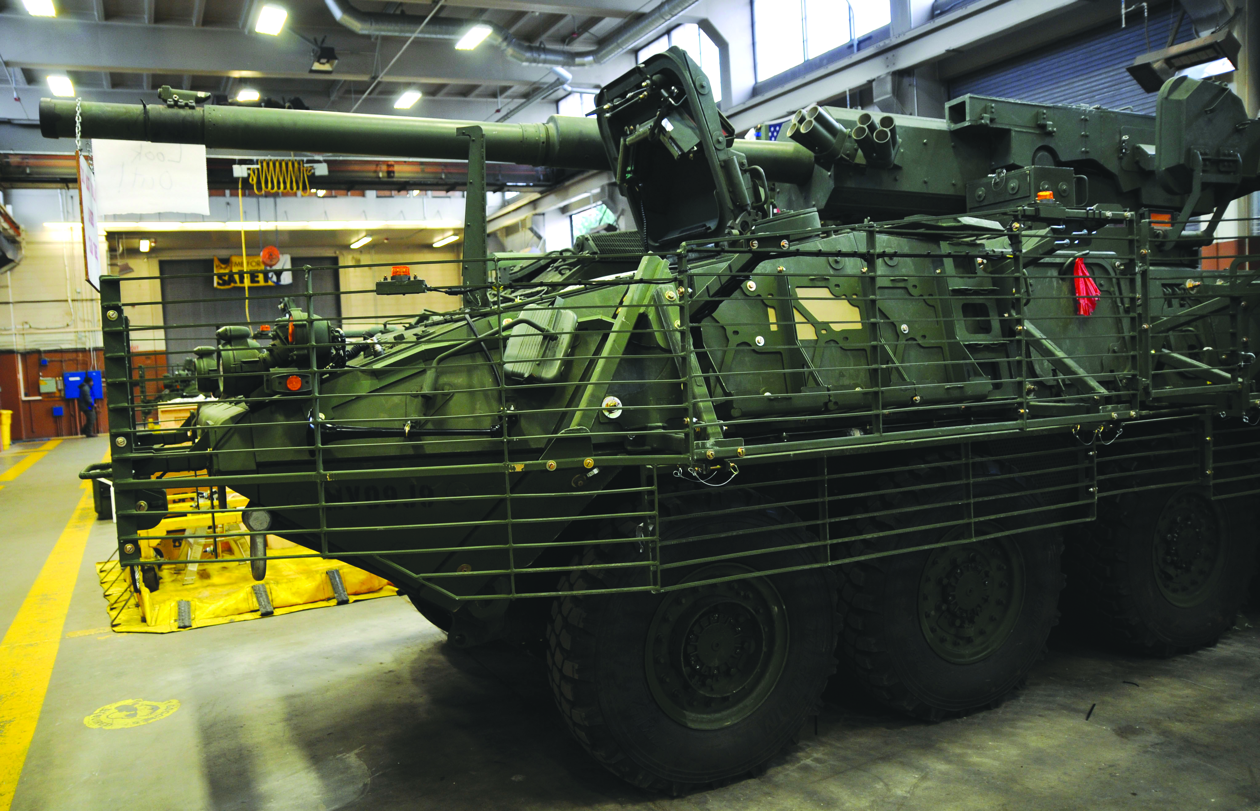 A green Stryker armored vehicle with slat armor is parked inside a maintenance bay or garage.